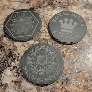 May include: Three dark gray slate coasters with engraved designs. One coaster reads "JEFFERSONIAN MEDICO LEGAL ANTHROPOLOGY UNIT". Another says "ROYAL DINER THE BEST BURGERS IN THE DISTRICT". The third says "DEPARTMENT OF JUSTICE FEDERAL BUREAU OF INVESTIGATION MAJOR CRIMES UNIT".