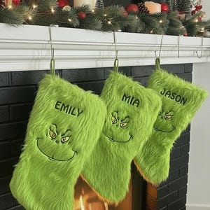 以下が含まれることがあります： 緑色のクリスマスストッキング3つ。グリンチの顔と、エミリー、ミア、ジェイソンと名前が書かれています。暖炉の棚に吊るされており、ガーランドやオーナメントで飾られています。