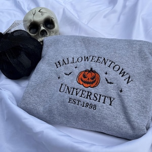 May include: A gray sweatshirt with the text "Halloweentown University" and a jack-o'-lantern graphic. The sweatshirt also features bats and stars. A black decorative hat and a skull are in the background.