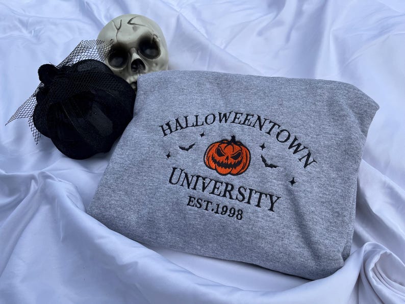 May include: A gray sweatshirt with the text "Halloweentown University" and a jack-o'-lantern graphic. The sweatshirt also features bats and stars. A black decorative hat and a skull are in the background.