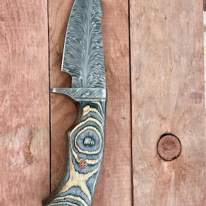 May include: A Damascus steel knife with a patterned blade and a wooden handle. The handle features a blue and tan wood grain design with a small red accent. The knife has a unique shape with a notch near the blade's base.