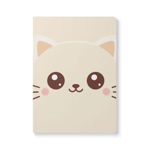 May include: A cream-colored notebook featuring a large, cartoon cat face on the cover. The cat has large brown eyes, pink cheeks, and a small black nose and whiskers. The notebook is rectangular and has a simple, clean design.