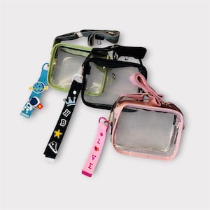May include: A collection of clear, rectangular crossbody bags in green, black, and pink. Each bag has a clear front panel and a colored trim. The bags are accessorized with colorful straps and charms, including a blue astronaut charm and a black strap with a crown.