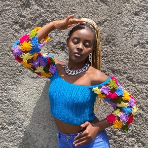May include: A blue off-the-shoulder crochet top with colorful floral sleeves. The sleeves feature a mix of yellow, pink, purple, and blue crochet flowers. The top is paired with light blue jeans.