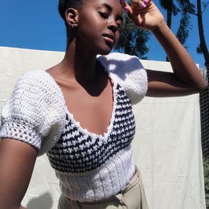 May include: A white and black crochet top with short, puffy sleeves. The top features a V-neck design with a black and white pattern on the bodice. The sleeves and lower portion of the top are white.