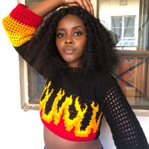 May include: A black crochet top with a flame design in red, yellow, and orange. The sleeves are long and open-weave, with red, yellow, and orange cuffs. The top is a cropped style.