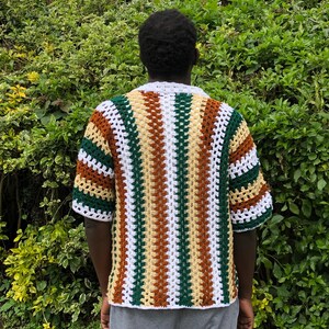 May include: A short-sleeved, crochet shirt in a striped pattern of white, brown, green, and yellow. The shirt has a white collar and is worn over grey sweatpants. The background is a lush green bush.