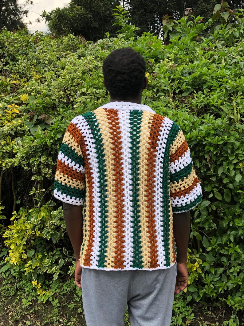 May include: A short-sleeved, crochet shirt in a striped pattern of white, brown, green, and yellow. The shirt has a white collar and is worn over grey sweatpants. The background is a lush green bush.