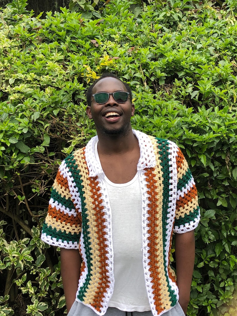 May include: A short-sleeved, open-front crochet shirt in a patchwork design. The shirt features alternating stripes of white, brown, green, and yellow. The collar is white, and the shirt is worn over a white undershirt.