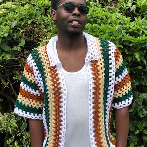 May include: A multi-coloured crochet shirt with short sleeves. The shirt has a striped pattern in brown, green, white, and tan. The shirt is open at the front and has a collar. The person is wearing sunglasses and a white t-shirt underneath.