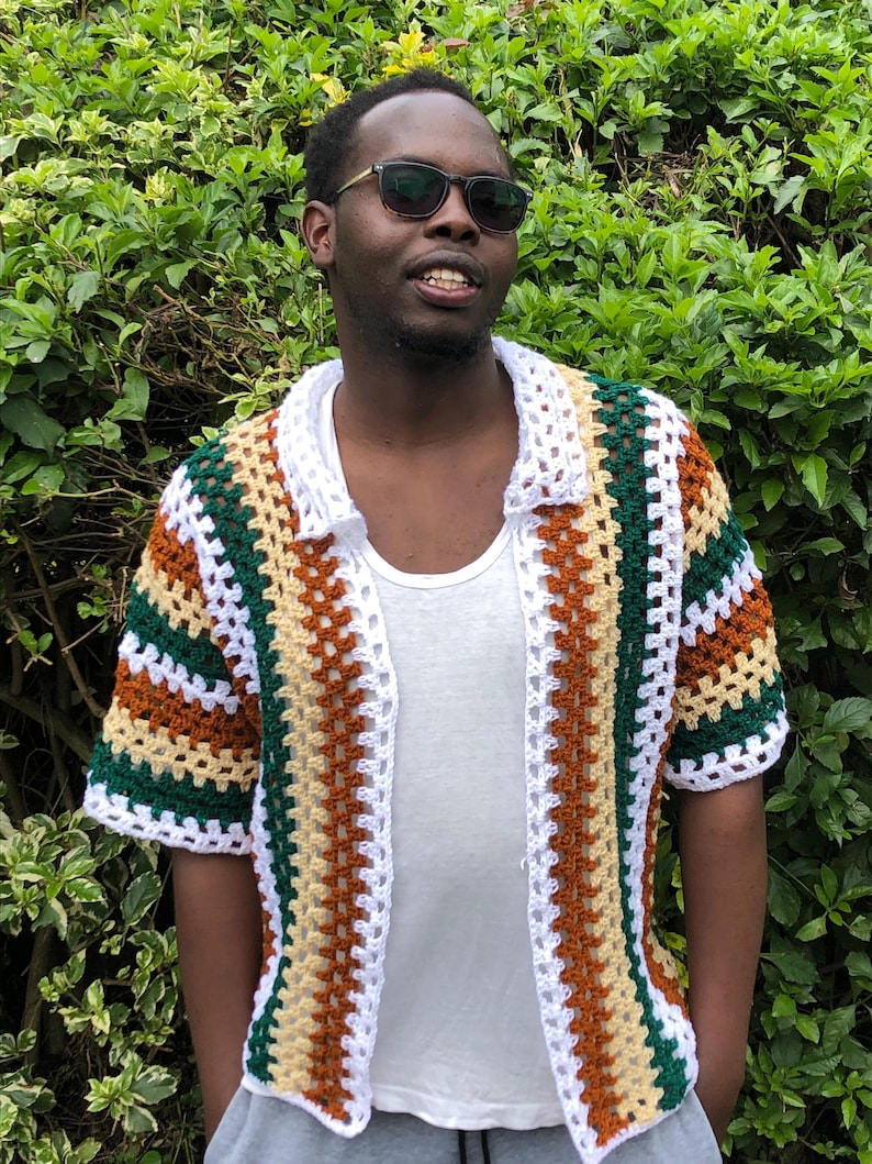 May include: A multi-coloured crochet shirt with short sleeves. The shirt has a striped pattern in brown, green, white, and tan. The shirt is open at the front and has a collar. The person is wearing sunglasses and a white t-shirt underneath.