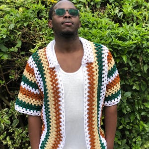 May include: A short-sleeved, open-front crochet shirt in a patchwork design. The shirt features alternating stripes of white, green, brown, and tan. The collar and edges are white. The shirt is worn over a white vest.