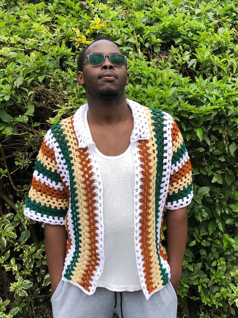 May include: A short-sleeved, open-front crochet shirt in a patchwork design. The shirt features alternating stripes of white, green, brown, and tan. The collar and edges are white. The shirt is worn over a white vest.