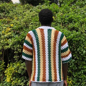May include: A short-sleeved crochet shirt in a striped pattern of white, green, brown, and tan. The shirt has a white collar and cuffs. The person is wearing grey sweatpants. The background is a green bush.