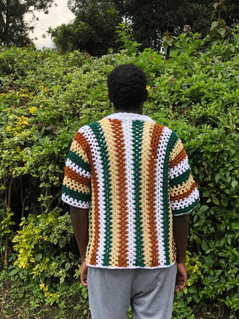 May include: A short-sleeved crochet shirt in a striped pattern of white, green, brown, and tan. The shirt has a white collar and cuffs. The person is wearing grey sweatpants. The background is a green bush.