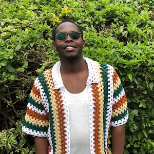 May include: A colourful, short-sleeved crochet shirt with an open front, featuring white, green, brown, and yellow stripes. The shirt has a collar and is worn over a white vest. The background is a green bush.