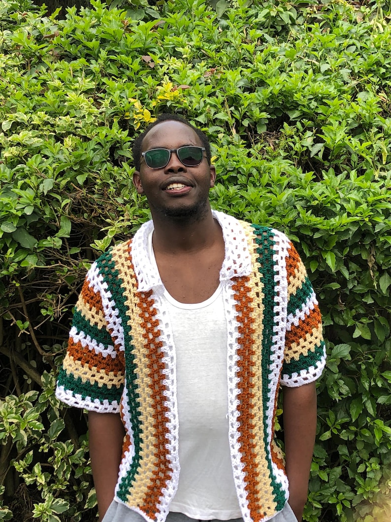 May include: A colourful, short-sleeved crochet shirt with an open front, featuring white, green, brown, and yellow stripes. The shirt has a collar and is worn over a white vest. The background is a green bush.