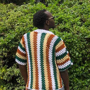 May include: A short-sleeved, open-weave crochet shirt. The shirt features vertical stripes in white, green, brown, and yellow. The collar and sleeve cuffs are white. The shirt is worn over a grey garment.