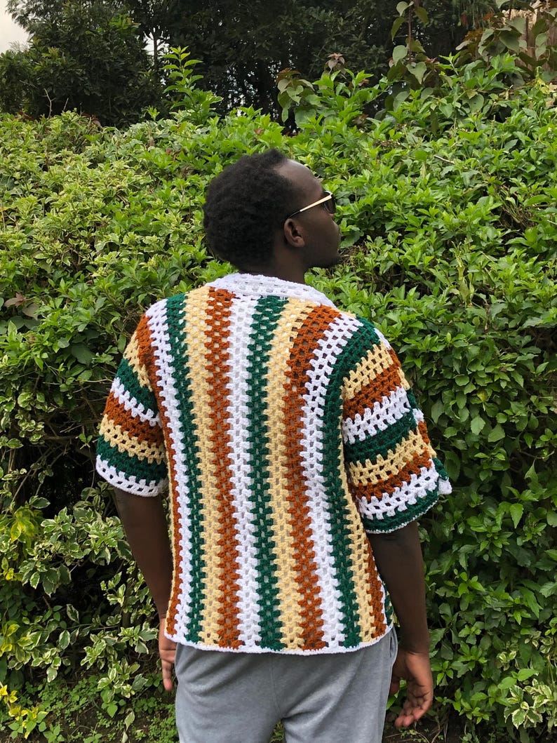 May include: A short-sleeved, open-weave crochet shirt. The shirt features vertical stripes in white, green, brown, and yellow. The collar and sleeve cuffs are white. The shirt is worn over a grey garment.