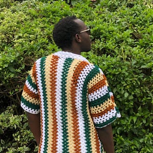 May include: A short-sleeved, open-weave crochet shirt with a striped design. The shirt has stripes of white, green, brown, and yellow. The collar and sleeve edges are white. The shirt is worn over a dark shirt.
