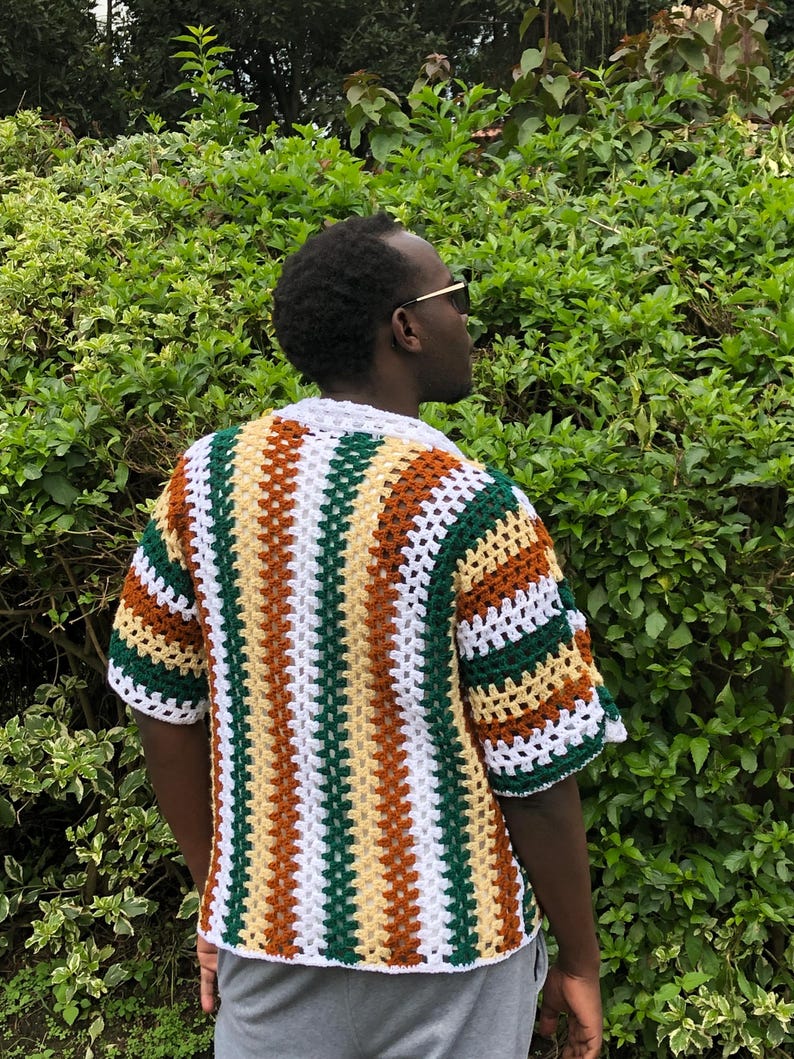 May include: A short-sleeved, open-weave crochet shirt with a striped design. The shirt has stripes of white, green, brown, and yellow. The collar and sleeve edges are white. The shirt is worn over a dark shirt.