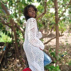 May include: A white, open-weave crochet cardigan. The long, flowing garment is worn over blue jeans and red sandals. The cardigan's texture and design are visible, with a focus on the open, airy weave. The background is a natural outdoor setting.