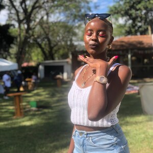 Puede incluir: Un top corto de crochet blanco combinado con vaqueros de mezclilla azul claro. La persona lleva un reloj de oro, un collar y pendientes de aro. Gafas de sol en la cabeza. El fondo presenta hierba verde y árboles.