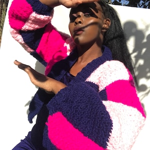 May include: A vibrant, patchwork crochet cardigan in navy blue, hot pink, and pale pink. The cardigan has large, voluminous sleeves and a V-neck. The handmade garment is a striking, statement piece, perfect for adding a pop of colour to any outfit.