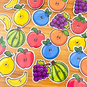 May include: A collection of colorful fruit stickers, including bananas, lemons, strawberries, blueberries, grapes, oranges, apples, and watermelons. Each sticker features a smiling face, adding a playful touch to the design. The stickers are arranged on a wooden surface.