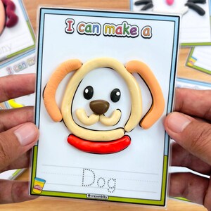 May include: A colorful card with a dog design made of clay. The card reads "I can make a Dog" with the text in a playful font. The dog has a beige face, brown nose, and orange ears. The card is held by a person.