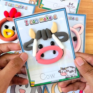 Puede incluir: Una tarjeta de actividad Play-Doh con una cara de vaca hecha de arcilla colorida. La tarjeta tiene el texto "I can make a" en la parte superior y "Cow" en la parte inferior. Otras tarjetas con caras de animales son visibles en el fondo.