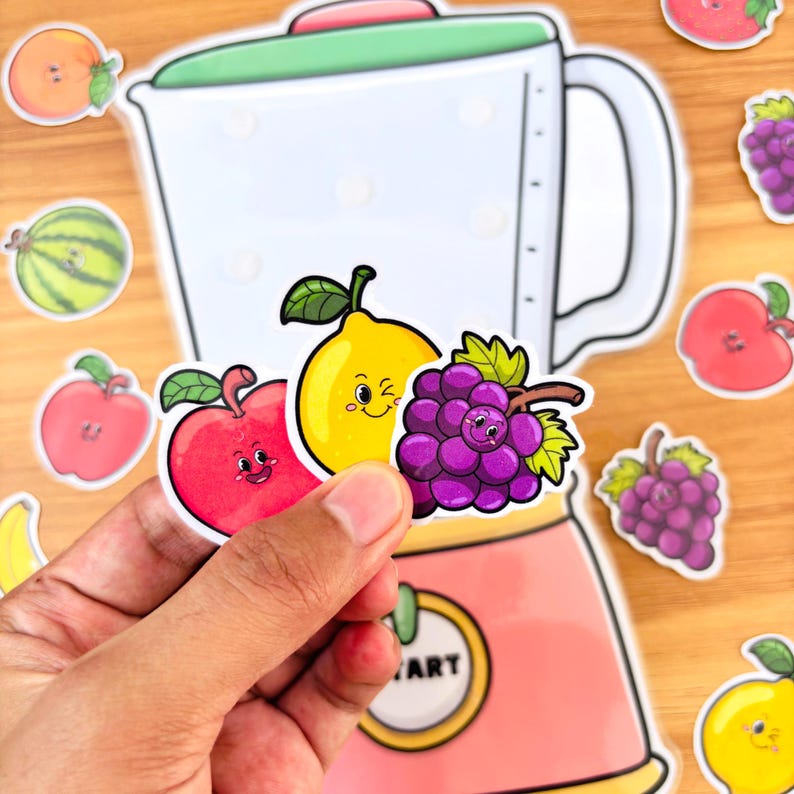 May include: A hand holding stickers of a red apple, yellow lemon, and purple grapes with smiling faces. A blender and other fruit stickers are in the background. The blender is white with a green lid and pink base.