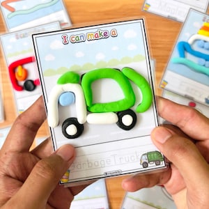 Op de afbeelding: Een kaart met de tekst "I can make a" en een klei vuilniswagen. De vrachtwagen is groen, wit en zwart, met een blauw accent. Andere kaarten met klei creaties staan op de achtergrond.