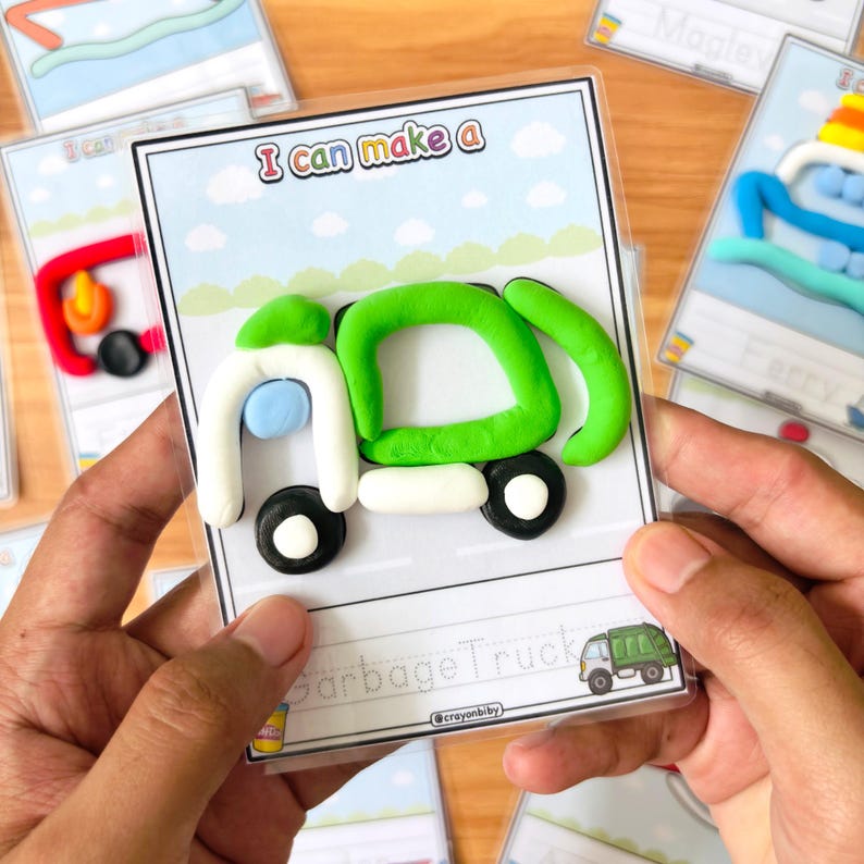 Op de afbeelding: Een kaart met de tekst "I can make a" en een klei vuilniswagen. De vrachtwagen is groen, wit en zwart, met een blauw accent. Andere kaarten met klei creaties staan op de achtergrond.