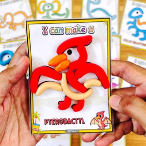 Puede incluir: Un Pterod&aacute;ctilo rojo y naranja hecho de plastilina en una tarjeta blanca con el texto "I can make a Pterodactyl". La tarjeta tiene un borde amarillo y una ilustraci&oacute;n de dinosaurio de dibujos animados. Otras tarjetas de dinosaurios est&aacute;n en el fondo.