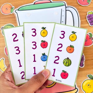 May include: Three white cards with green borders are held in a hand, each displaying numbers and colorful fruit illustrations. A blender sticker and fruit stickers surround the cards. The fruit includes lemons, apples, blueberries, and watermelon.