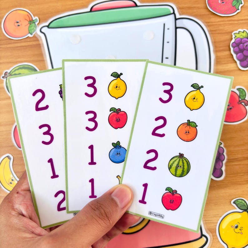 May include: Three white cards with green borders are held in a hand, each displaying numbers and colorful fruit illustrations. A blender sticker and fruit stickers surround the cards. The fruit includes lemons, apples, blueberries, and watermelon.