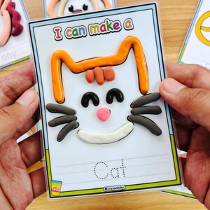 May include: A card with a cat face made of orange, black, pink, and white clay. The card reads "I can make a" at the top and "Cat" at the bottom. The card is held by hands.