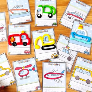 Op de afbeelding: Een verzameling activiteitkaarten met transportthema. Elke kaart toont een lijntekening van een voertuig, zoals een brandweerwagen, helikopter of schoolbus, met een bijpassend kleimodel. De kaarten liggen op een houten oppervlak.