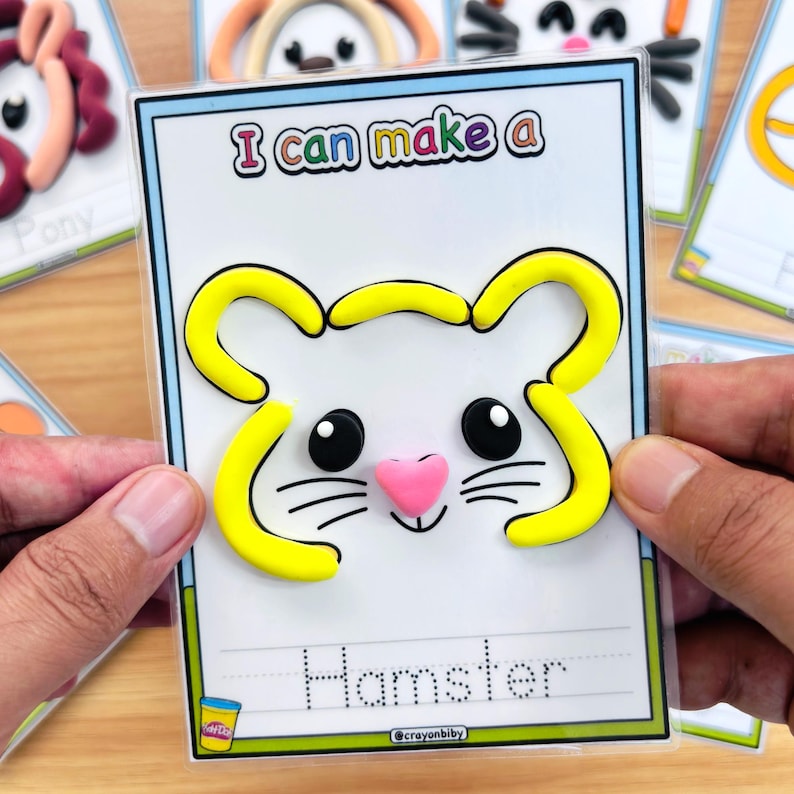 May include: A card with a hamster design made of yellow and pink clay. The card reads "I can make a" at the top and "Hamster" at the bottom. The card is held by a person's hands.