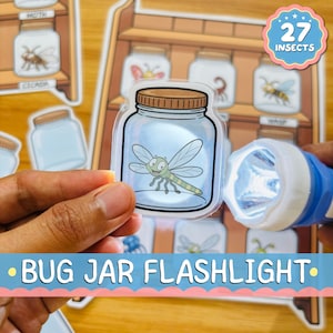 May include: A clear plastic sticker shaped like a mason jar with a brown lid, featuring a cartoon dragonfly inside. A blue and white flashlight shines light through the sticker, which is held in front of a printed sheet showing various insects.