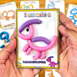 Puede incluir: Un dinosaurio parasaurolophus rosa y morado hecho de plastilina Play-Doh se muestra en una funda de pl&aacute;stico transparente. El texto "I can make a" est&aacute; en la parte superior y "PARASAUROLOPHUS" en la inferior. Hay otras tarjetas de dinosaurios en el fondo.