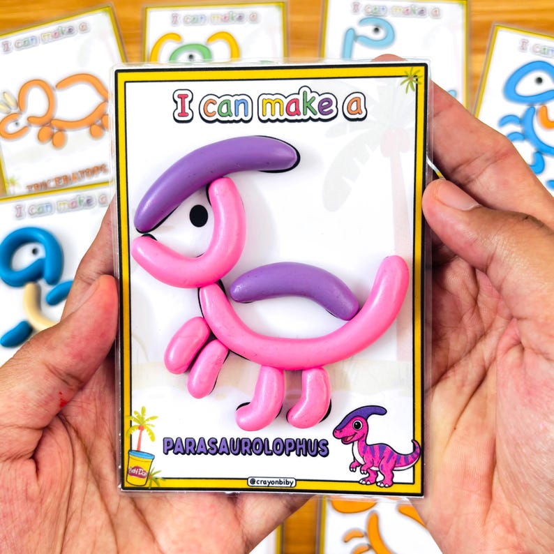 Puede incluir: Un dinosaurio parasaurolophus rosa y morado hecho de plastilina Play-Doh se muestra en una funda de pl&aacute;stico transparente. El texto "I can make a" est&aacute; en la parte superior y "PARASAUROLOPHUS" en la inferior. Hay otras tarjetas de dinosaurios en el fondo.