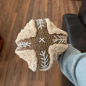 May include: A handmade, crocheted pillow with a cross design. The centre is brown with white stitching in a cross pattern. The edges are a soft, beige material. A unique, decorative item.
