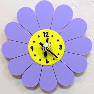 Peut inclure: Une horloge murale en forme de fleur avec douze pétales violets arrondis entourant un cadran circulaire jaune vif. L'horloge présente des chiffres noirs aux positions 12, 3, 6 et 9, des aiguilles noires et une petite figurine d'abeille au centre.