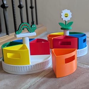 May include: Brightly coloured, segmented storage containers with a rotating design. Each container is divided into sections of yellow, red, blue, and green. One has a green caterpillar topper, and another has a white flower topper. The containers sit on a white base.