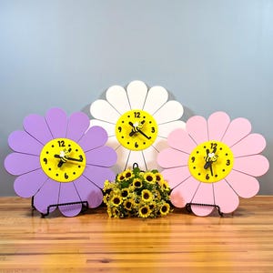 Peut inclure: Trois horloges murales en forme de fleur, de couleur violette, blanche et rose, avec un centre jaune vif, des chiffres noirs et une petite icône d'abeille sur les aiguilles. Un bouquet de petits tournesols artificiels est placé devant.