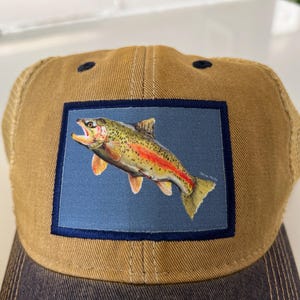 Puede incluir: Gorra de camionero marrón con parte trasera de malla y visera azul oscuro. La gorra presenta un parche rectangular con una colorida ilustración de un pez trucha sobre un fondo azul, enmarcado por un borde azul oscuro.