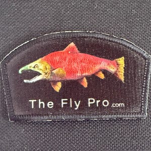 May include: Black patch featuring a red and yellow fish design with an open mouth and green accents. The text "The Fly Pro.com" is printed in white. The patch has a white stitched border.