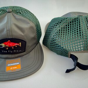May include: Two olive green baseball caps with mesh backs. One cap features a patch with a red fish and the text "The Fly Pro.com". The other cap is turned to show the mesh back and adjustable strap. The caps have a curved brim.